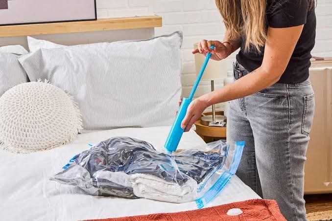 FB_IMG_1769212727664 Woman using a blue manual pump to remove air from a clear vacuum storage bag filled with clothes on a white bed.